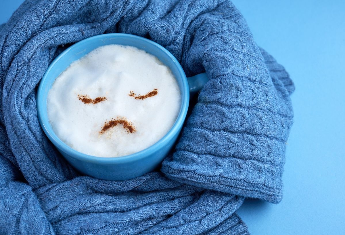 Kék háttéren kék kötött pulóver ujjába bugyolált kék bögre, amiben cappuccinó van, a tetejére kakaóporból szomorú arcot szórtak