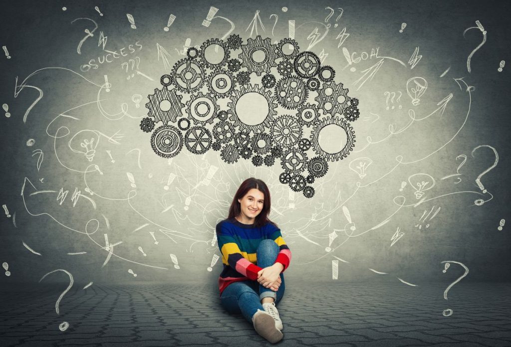 Nő színes pulóverben ül a szürke fal előtt agy alakban fogaskerekek, nyilak és kérdések körülötte