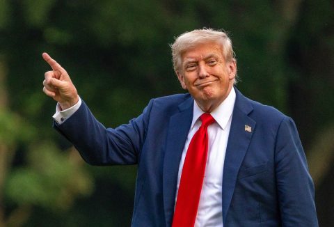 Donald Trump mosolyog grimaszolva kék öltönyben, piros nyakkendőben, mutatóujjával felfelé mutat