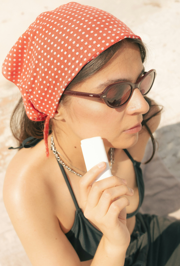 Nő napszemüvegben, piros alapon fehér pöttyös kendőben fényvédő stifttel keni az arcát a strandon