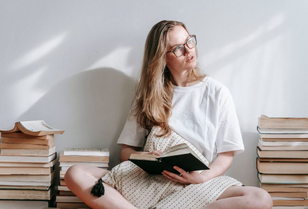 Hosszú, szőkehajú, szemüveges nő könyvvel a kezében ül fehér pólóban könyvkupacok között