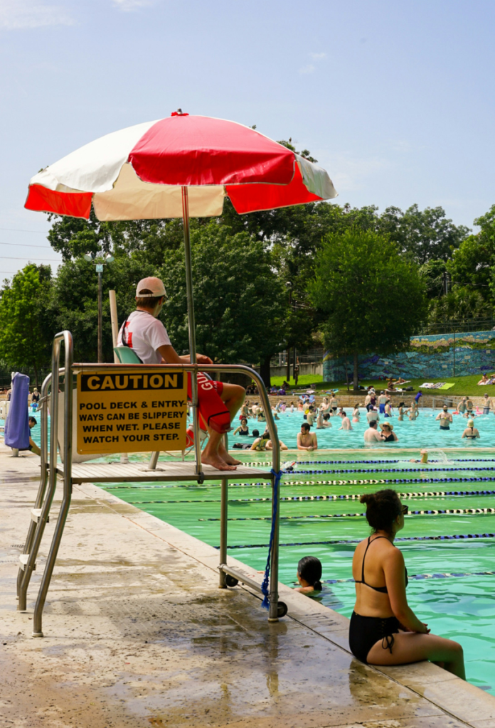 strandolas-mennyire-veszelyes-megelozes | Marie Claire Nyilvános strandon úszómester a medence szélén magas széken ül fehér-piros napernyő alatt, bikins nő lógatja a lábát a medencébe
