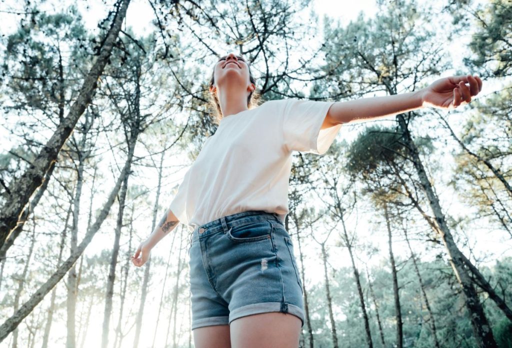 felszabadito-asztrologiai-esemenyek-2025-augusztus | Marie Claire Farmer rövidnadrágos, fehér pólós nő alulról fotózva széttárt karral fák között