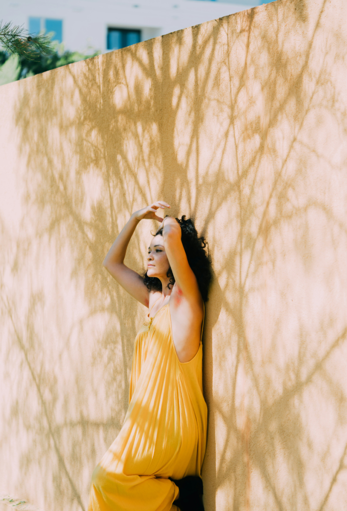 Fehér fal egy fa árnyékaival, amihez egy szép, sötét hajú, sárga nyári ruhás nő támaszkodik
