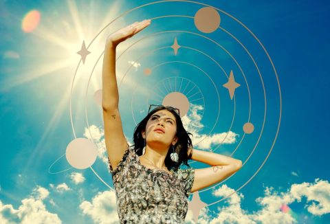 Napsütéses égbolt és asztrológiai szimbólumok előtt fekete hajú nő néz az ég felé feltartott kézzel árnyékolva a szemét