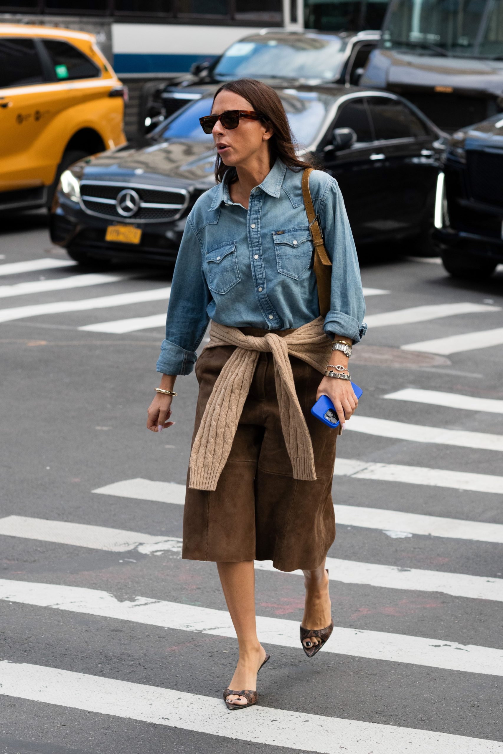 Egy nő kék farmeringben, barna velúr rövidnadrágban, barna pulóverben és barna szandálban.