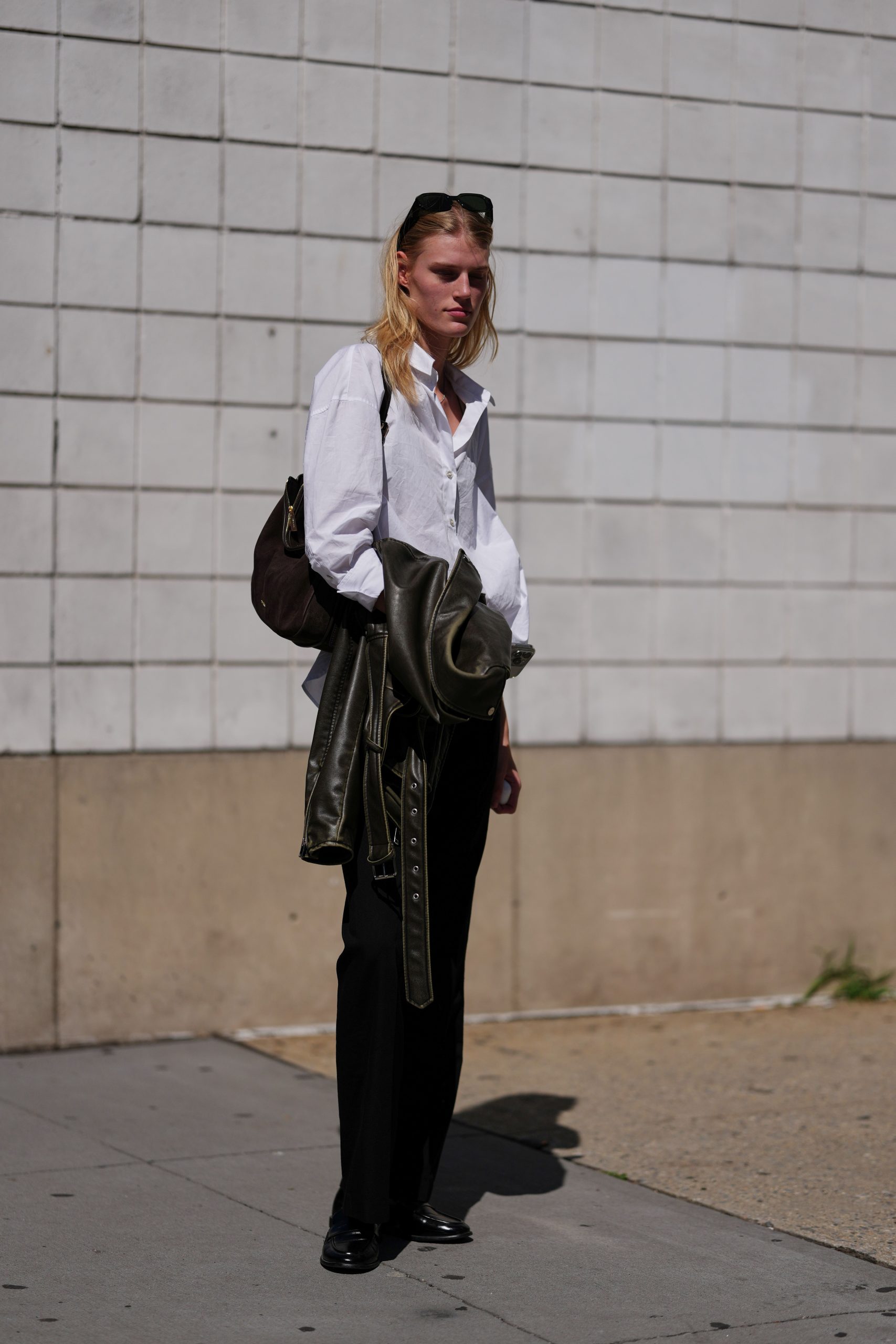 Egy nőke nő fehér, oversize pamutinget visel széles gallérral és gombos mandzsettával, karján egy sötét olívazöld bőrből készült motorosdzseki