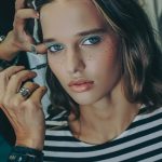 Szép, fiatal modellt sminkelnek a KATA Szegedi divatshow-ra, csíkos pólóban, műszeplővel, kék szemsminkkel