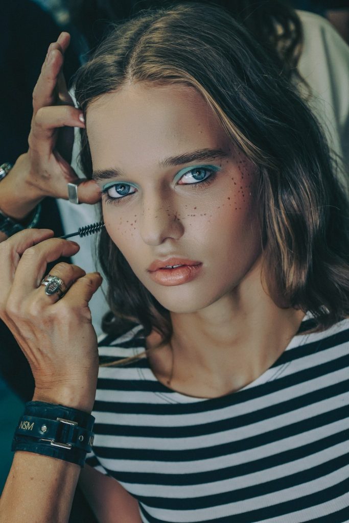 Szép, fiatal modellt sminkelnek a KATA Szegedi divatshow-ra, csíkos pólóban, műszeplővel, kék szemsminkkel