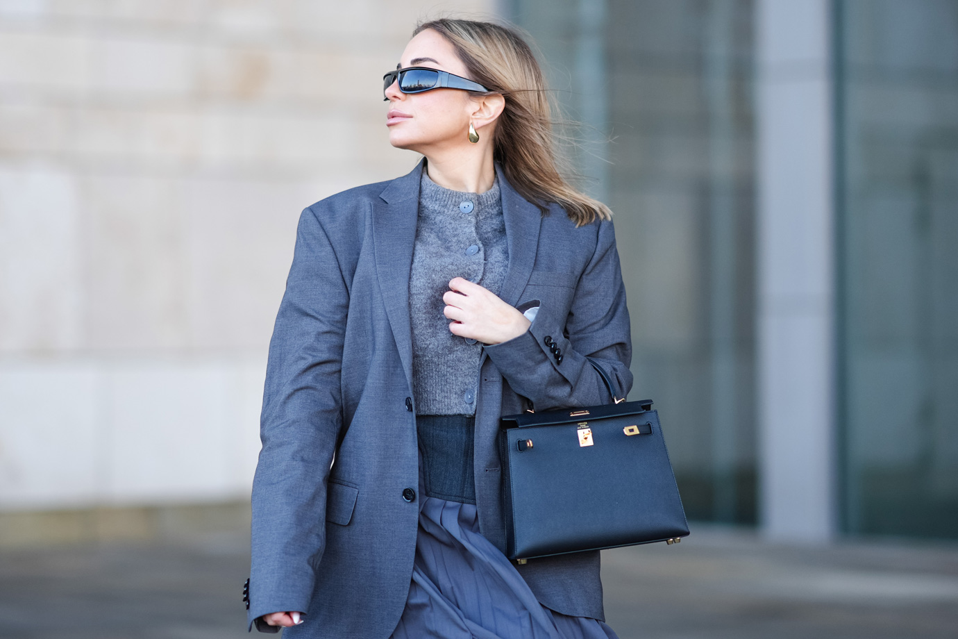 Egy napszemüveges nő nagyméretű blézert visel és táska van a karján