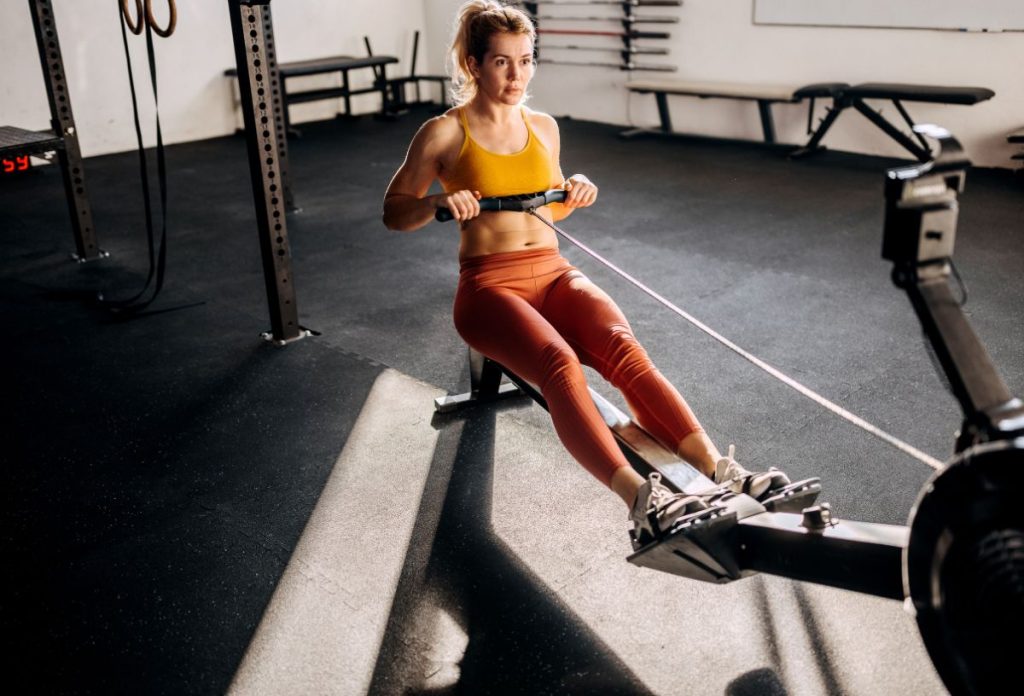 Nő sárga sportmelltartóban és rozsdavörös leggings-ben evezőpadon edz az edzőteremben