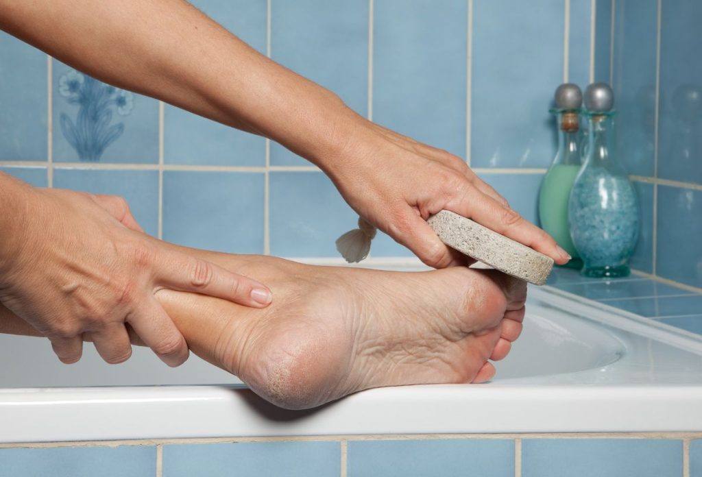 Nő a fürdőkád szélén habkővel távolítja el a bőrkeményedést a talpáról