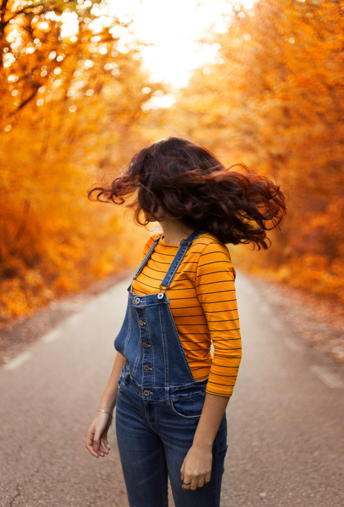 Barna hajú nő fordul a kamera előtt farmer kertésznadrágban egy úton, amit sárga lombú fák szegélyeznek