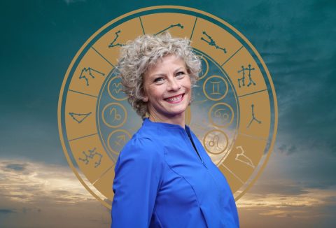 Német színésznő kék ruhában, mosolyogva, szőke, göndör rövid hajjal, mögötte asztrológiai jegyek körben, kékesszürke égbolt