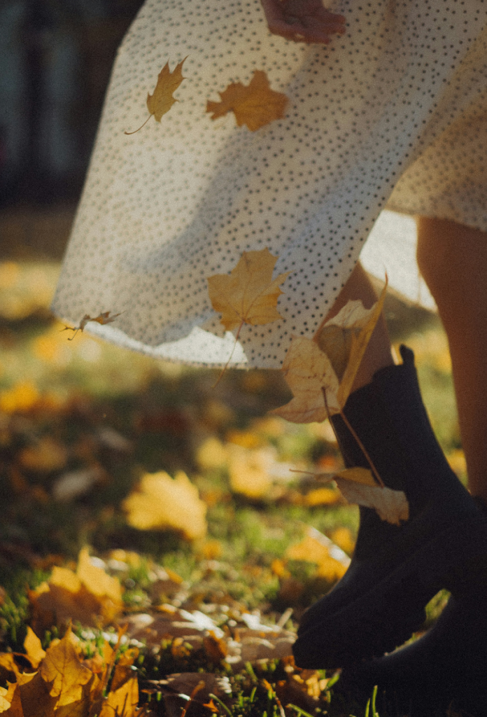 Őszi falevelek között sétáló nő lába fehér alapon fekete apró pöttyös, hosszú szoknyában, bokacsizmában