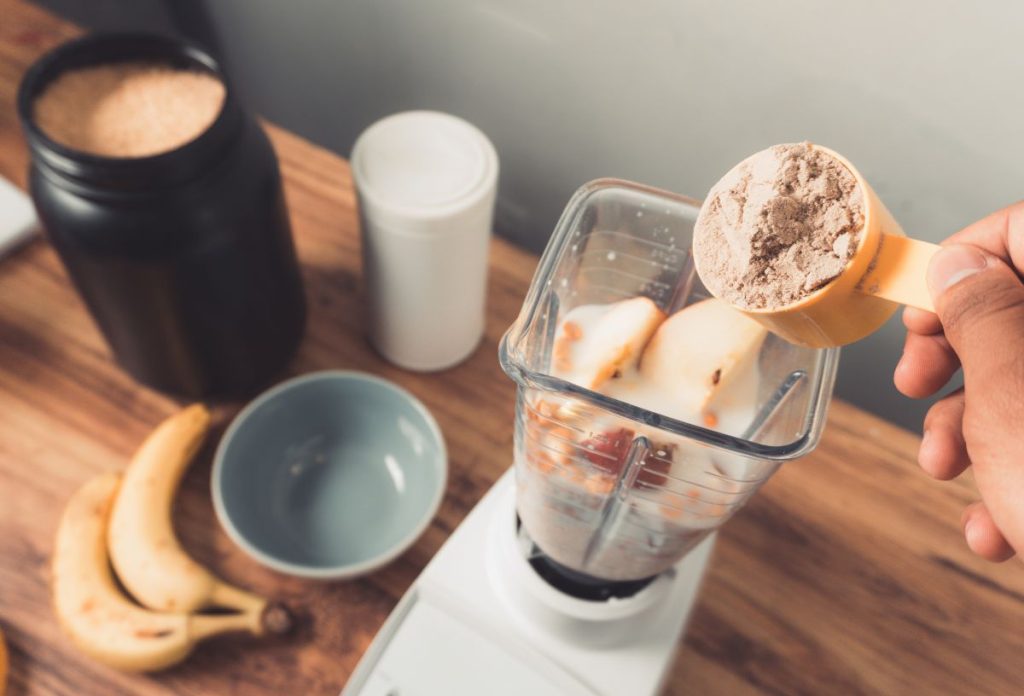 Egy kanálnyi kreatinport rak egy kéz turmixgépbe, mellette banán és a táplálékkiegészítő doboza