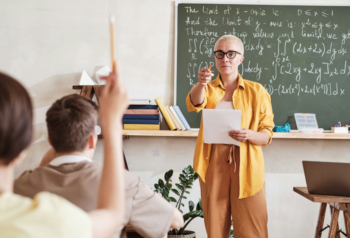 Rövid, szőke hajú tanár sárga ingben és barna nadrágban mutat az osztályban az egyik jelentkező diákra