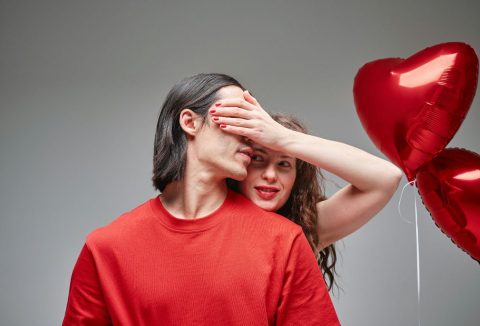 Nő piros rúzzsal és körömlakkal befogja egy hosszú hajú, piros pulóvert viselő férfi szemét, mellettük piros, szív alakú lufik