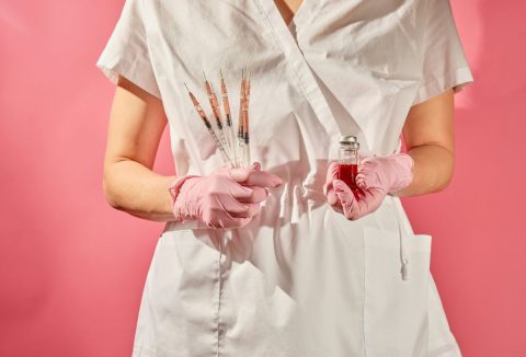 Minimalista képen, rózsaszín háttérrel ápolónő injekciós tűkkel és oltással a kezében