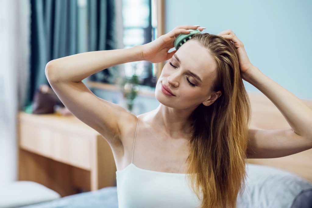 Fiatal, hosszú hajú nő a fejbőrét masszírozza egy eszközzel hajhullás ellen