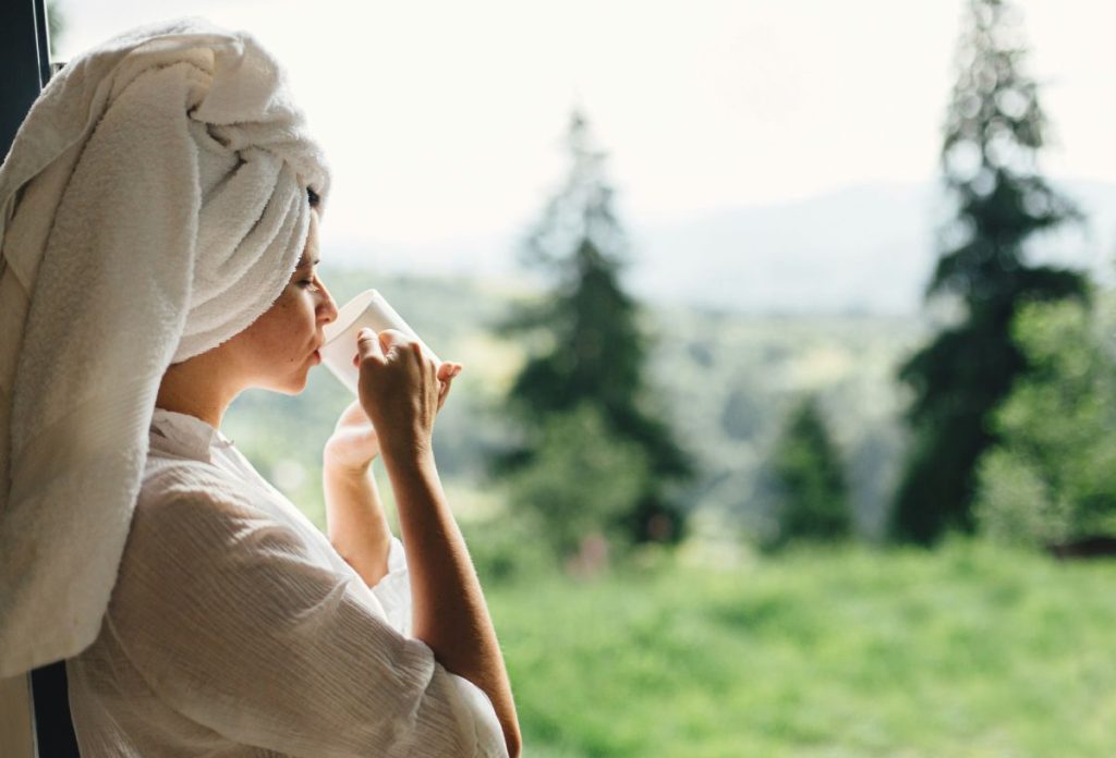 Nő fehér törölközőbe csavart hajjal és köntösben ablaknál állva fehér csészéből iszik