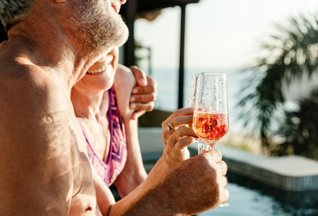 Idős pár medence partján rosé pezsgőt iszik, a férfi átöleli a nőt
