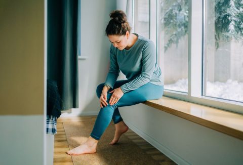 40 feletti nő ül az ablakpárkányon és a fájós térdét fogja