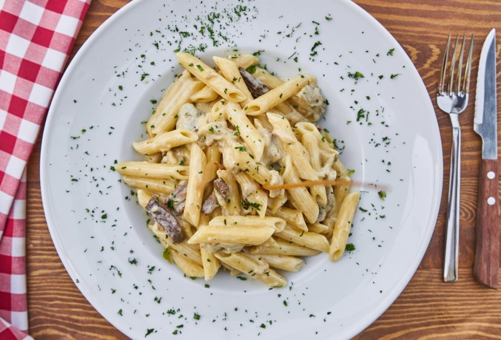 Fehér tányérban penne tészta fehér szósszal, zöldfűszerrel meghintve, mellette evőeszköz és piros-fehér kockás asztalkendő