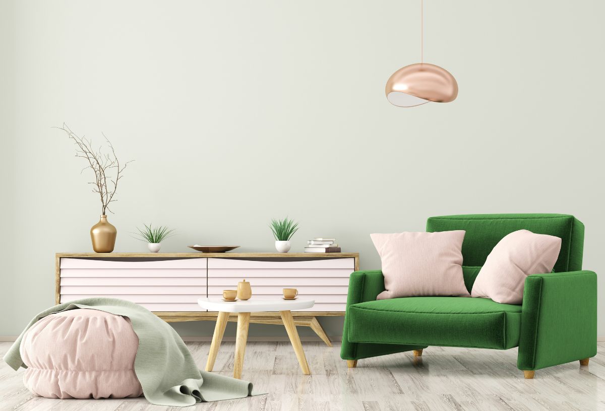 Vidám lakásbelső zöld fotellel, rózsaszín díszpárnákkal, puffal és komóddal. A fotel mögött króm rózsaszín állólámpa.