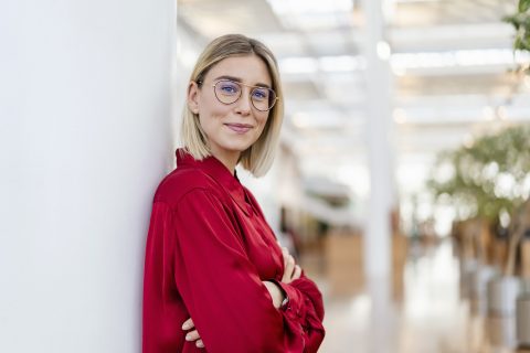 oszlopnak támaszkodó piros inges szemüveges nő a kamerába mosolyog
