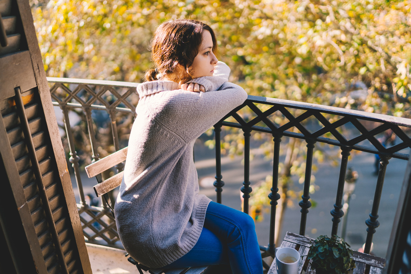 Egy nő az erkélyen a korlátnak dőlve szomorúan néz az őszi időben