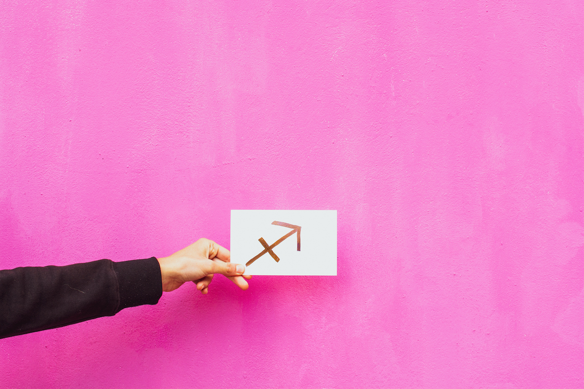 fekete pulóvert viselő kéz a Nyilas csillagjegy (íjász nyila) szimbólumát tartja rózsaszín háttér előtt