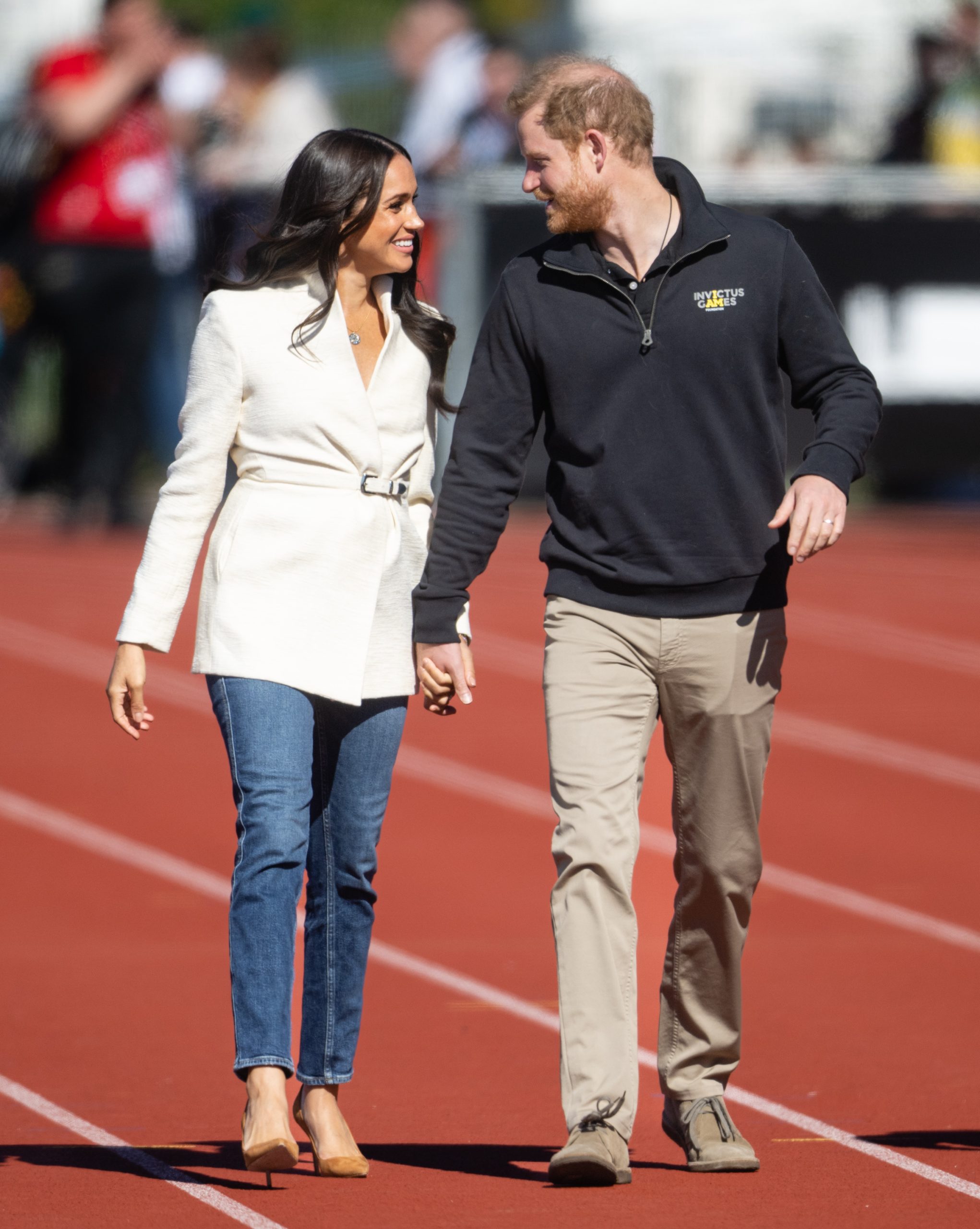 Meghan Markle fehér őszi kabátban, Harry herceg kezét fogja és egymásra mosolyognak