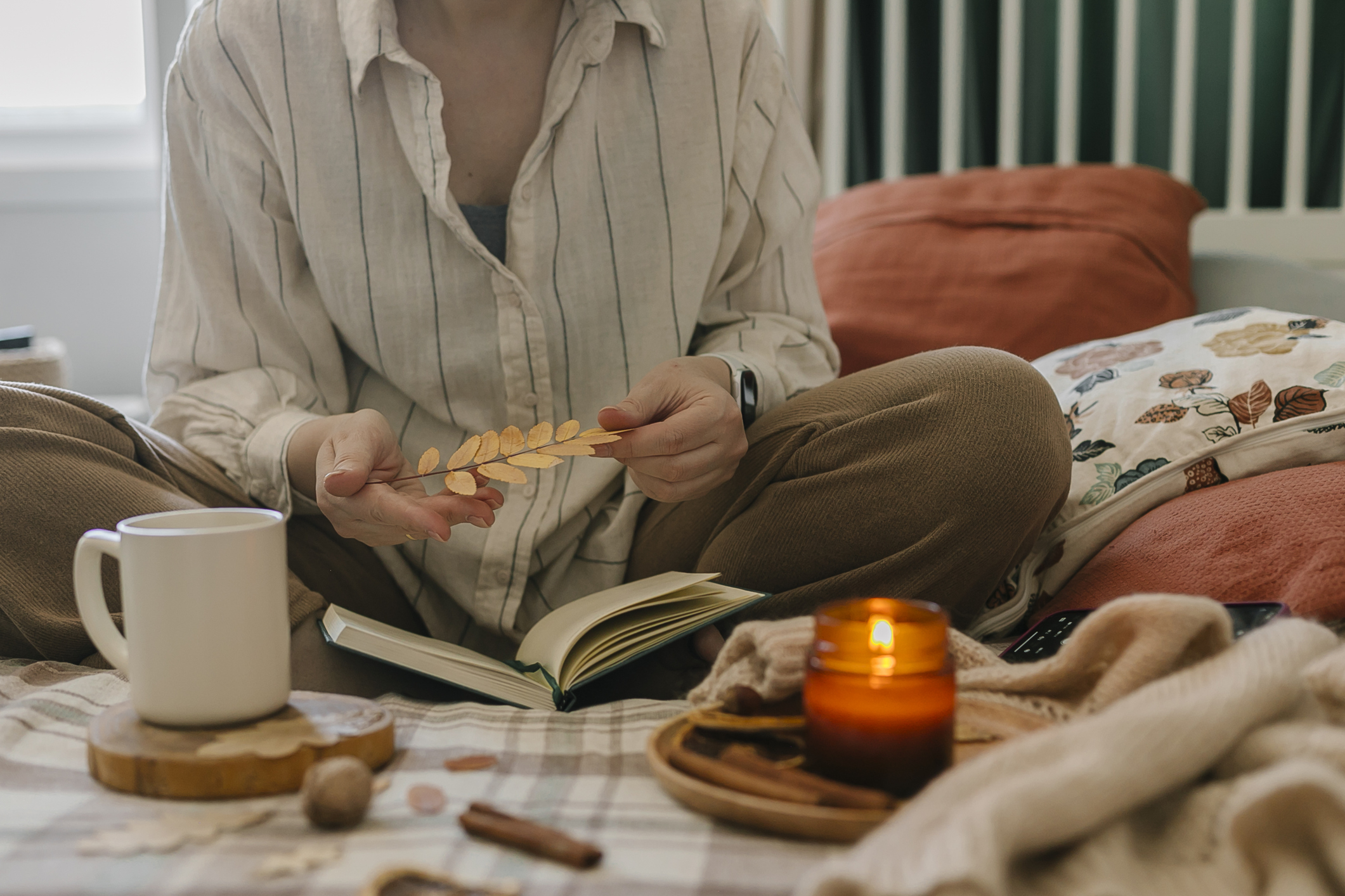 Nő ül az ágyon mécsessel, fahéjrudakkal, könyvvel és őszi falevéllel