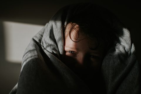 Egy gyerek kikukucskál a vastag, puha, szürke paplan alól. Arcát nagyrészt eltakarja az ágynemű és az árnyékok.