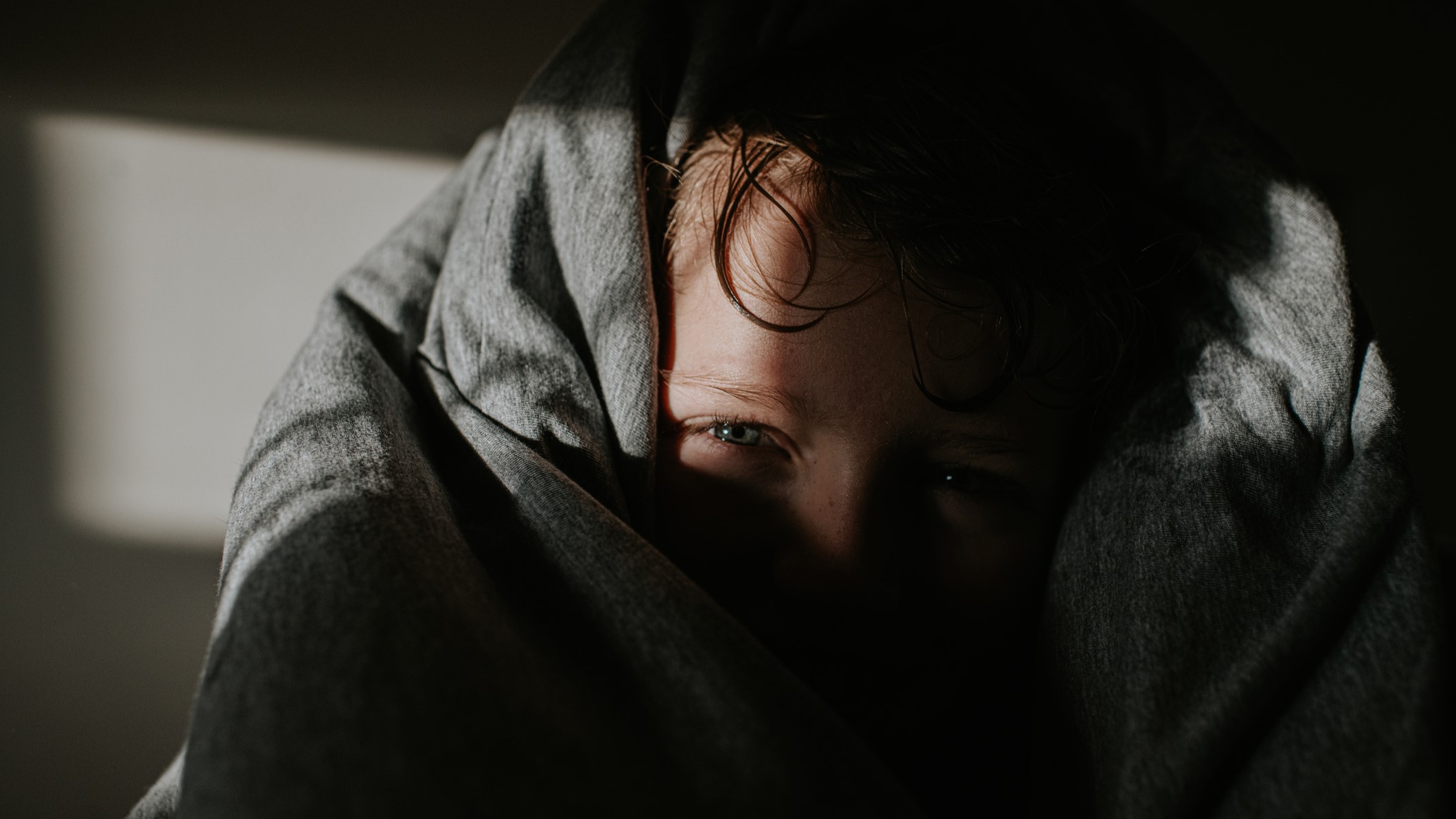 vilagszep-alapitvany-az-allami-gondozott-gyerekek-nehezsege | Marie Claire Egy gyerek kikukucskál a vastag, puha, szürke paplan alól. Arcát nagyrészt eltakarja az ágynemű és az árnyékok.