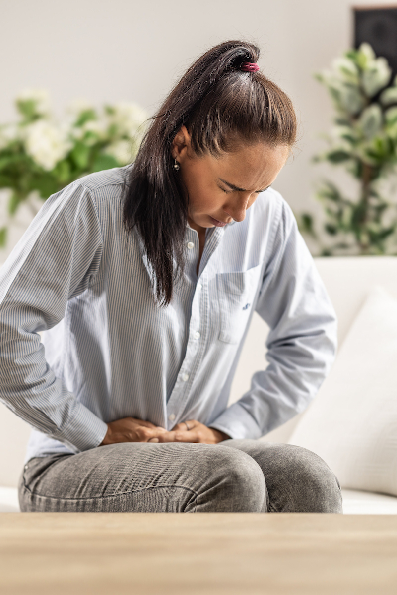 Egy fiatal nő otthoni ruhában ül a kanapén, mindkét kezével a hasát fogja, és előrehajol a fájdalom miatt.