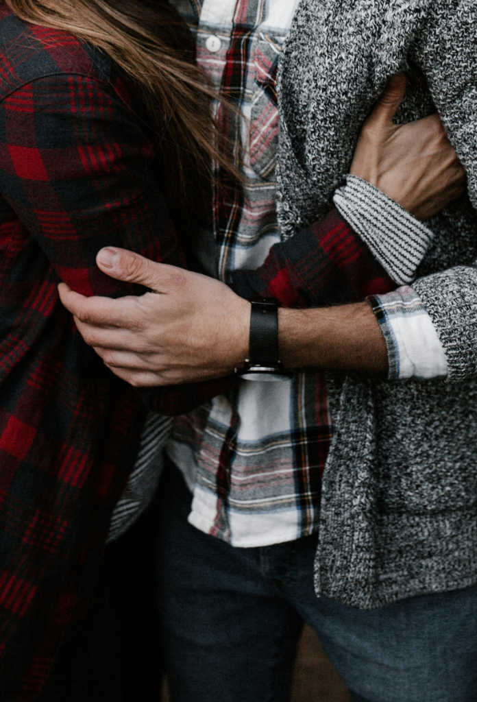 Közeli kép egy párról, a nő piros-fekete flanelingben a férfi hónalja alá dugja a kezét, a férfi a könyökénél karolja szürke pulóverben a nőt