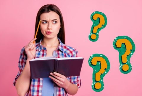 Fiatal nő erősen gondolkodik, ceruzáját a halántékához támasztja, kezében könyv, rózsaszín háttér előtt kockás ingben