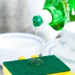 Zöld mosogatószert önt sárga gumikesztyűs kéz mosogatószivacsra