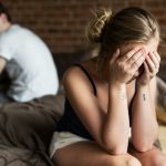 Szomorú nő ül az ágyon, háta mögött a férfi.