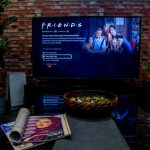 Képernyőn a Jóbarátok sorozat, előtte dohányzóasztalon magazin és snackek