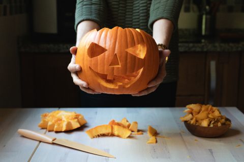 Egy nő egy ijesztő arcú halloweeni tököt tart a kezében, nem messze tőle kés és tökdarabok