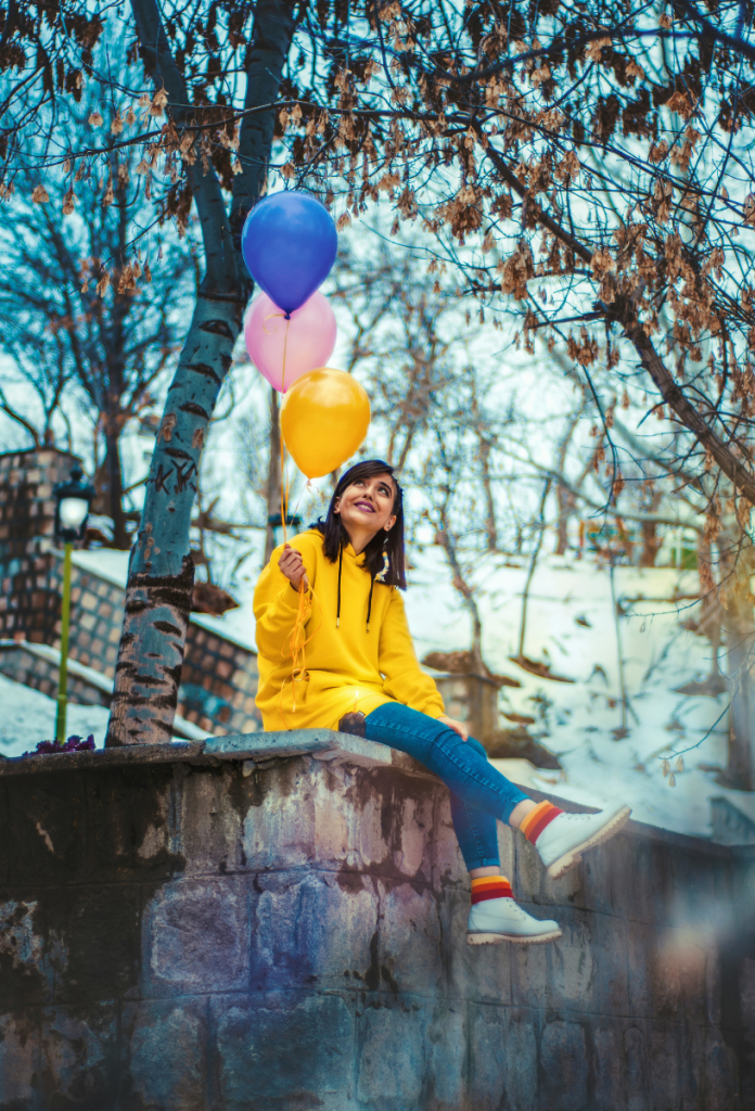 Barna hajú nő sárga esőkabátban, csíkos zokniban ül a fal tetején, kezében egy sárga, egy rózsaszín és egy kék lufi