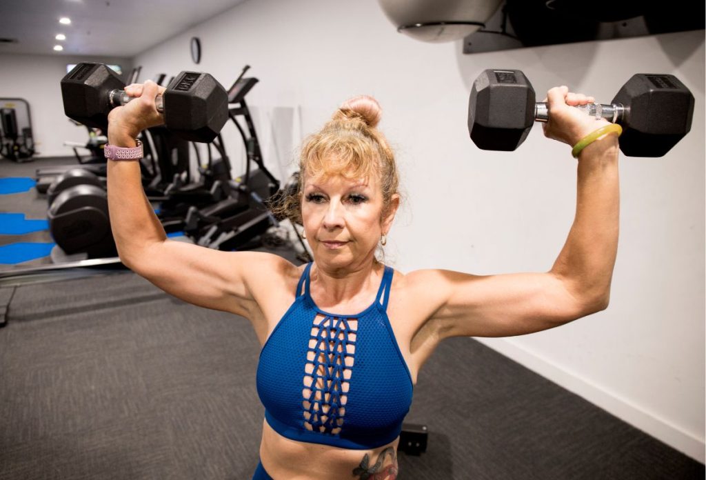 60 feletti nő edzőteremben súlyzókkal edzi a karját kék sportmelltartóban