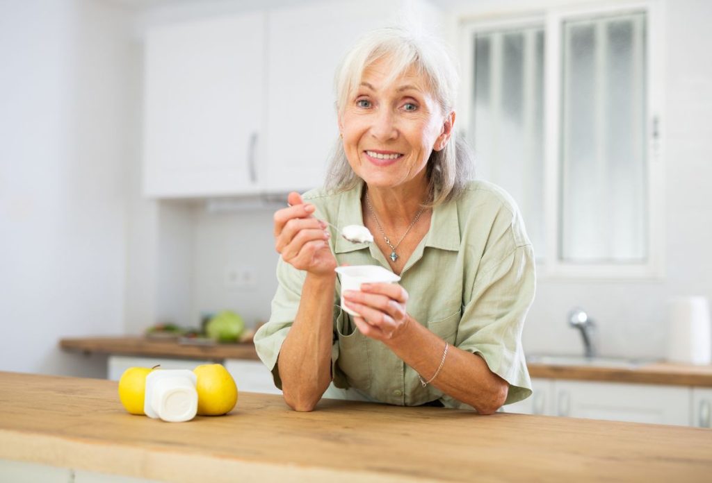 Idős, fehér hajú, csinos nő joghurtot kanalaz asztalra könyökölve