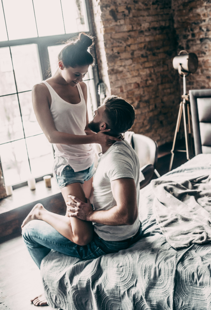 Szakállas férfi az ágy szélén ül, dekoratív fiatal nő fehér trikóban és farmer rövidnadrágban az ölében térdel és a szemébe néz, a kezével a vállát fogja