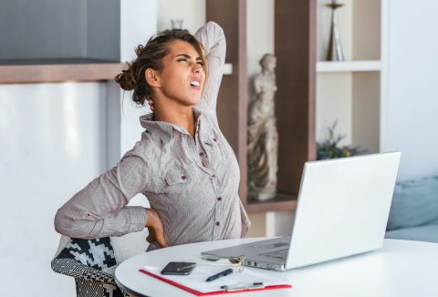 Fiatal nő grimaszolva nyújtózkodik, fájdalmasan fogja a vállát és nyakát az íróasztalnál ülve