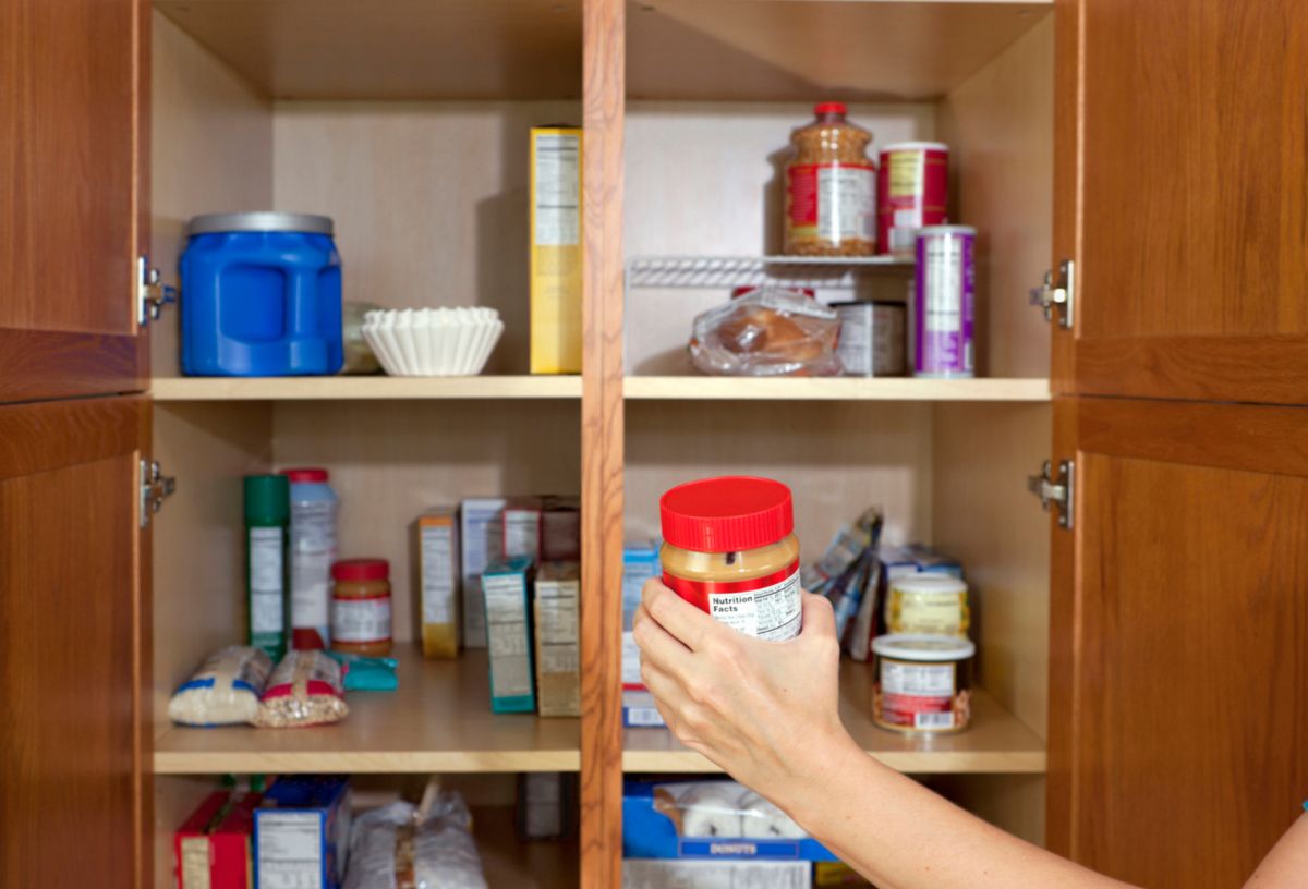 Nő pakol a kamraszekrényben, piros tetejű üveget tesz a polcra a dobozok és befőttek közé