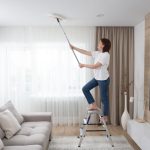 Nő nappali szobában szobalétrán állva seprűvel tisztítja a plafont
